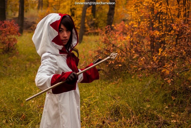 A person dressed in a white cloak with red accents holds a silver staff in a forest with autumn foliage.