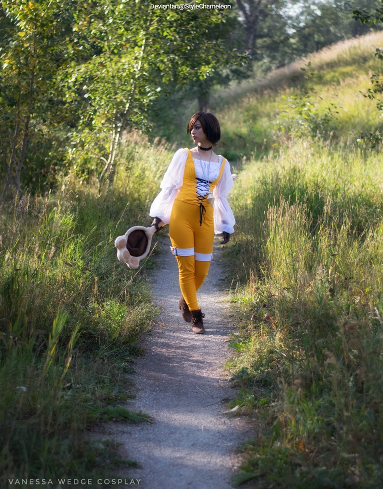 A person in a yellow outfit and white blouse walks along a path in a grassy area, holding a hat in one hand.