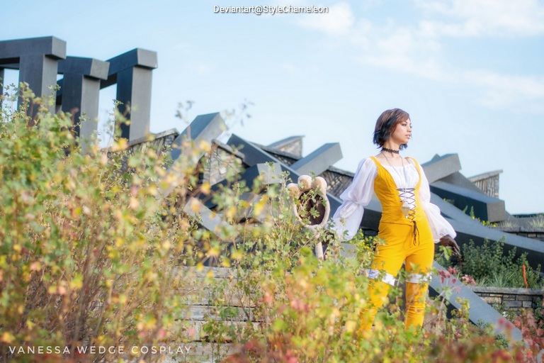 A person dressed in a yellow cosplay outfit stands amidst greenery, with an abstract architectural structure in the background.