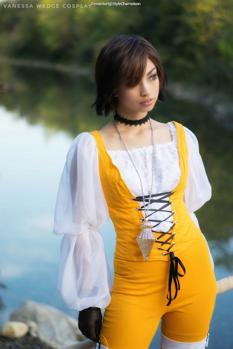 A person dressed in a yellow and white cosplay outfit, with a choker and pendant, standing near a body of water surrounded by greenery.