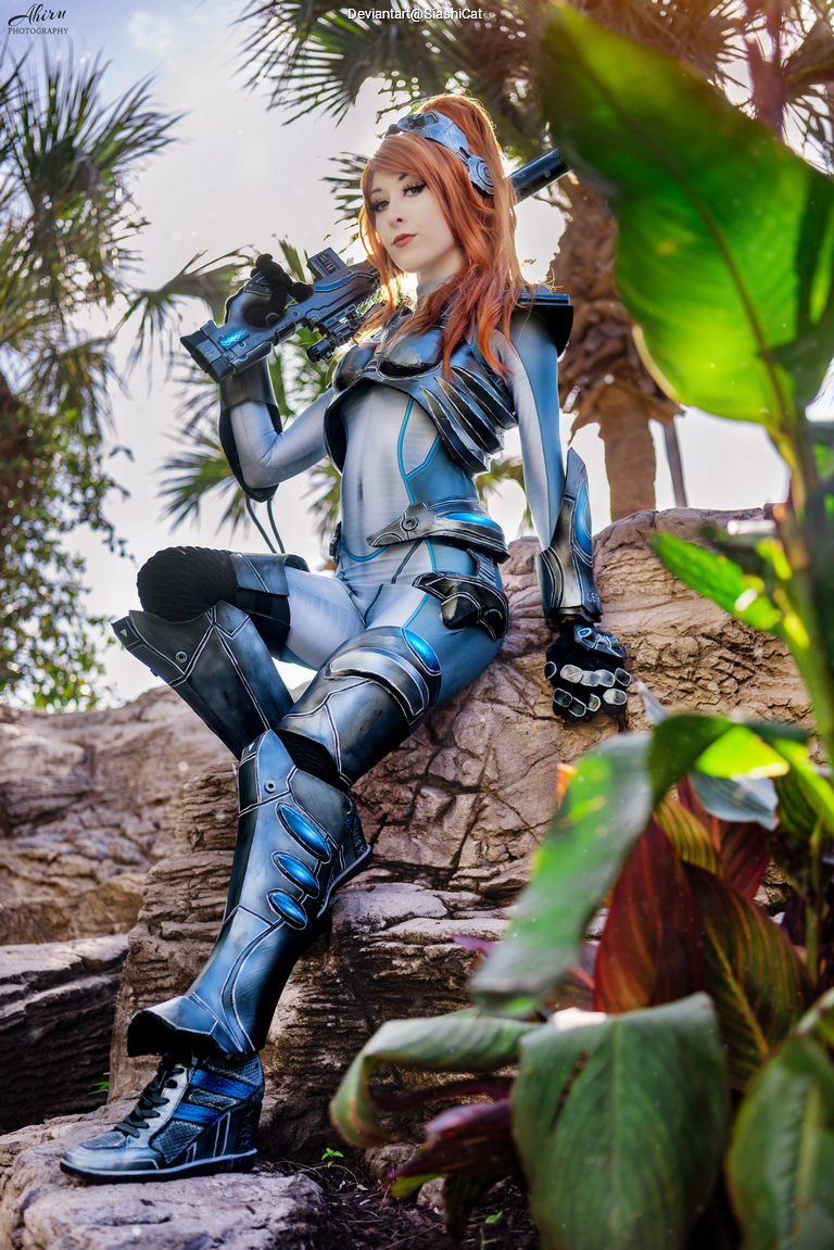 A person in a futuristic blue and silver costume holds a weapon while sitting on a rock formation surrounded by tropical plants.