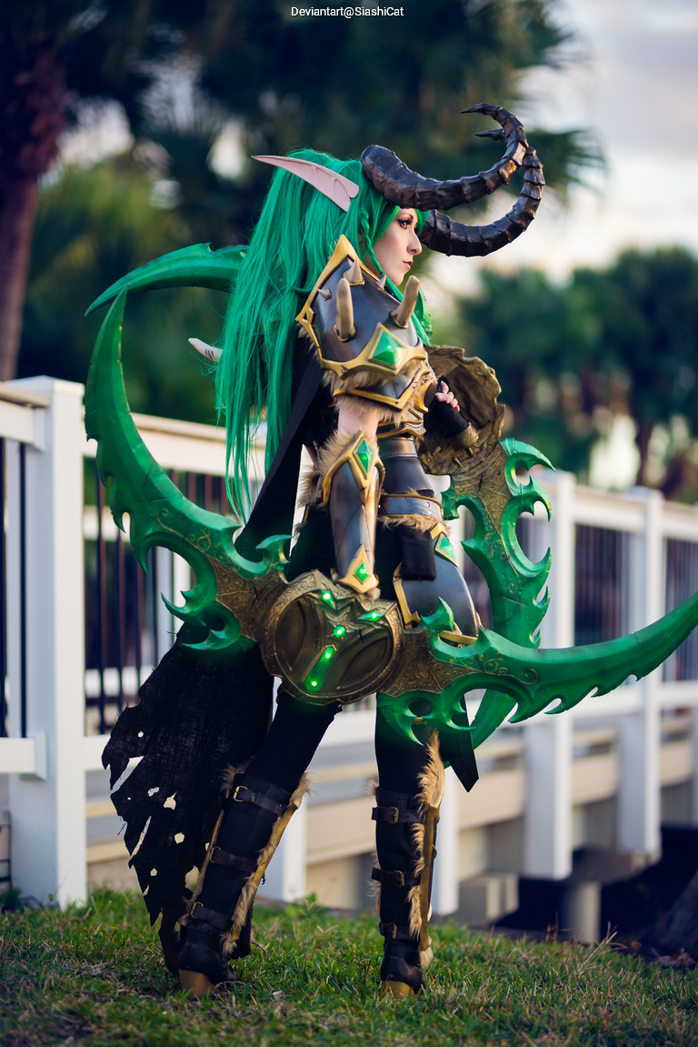 A person in detailed fantasy cosplay with long green hair and large horns, wielding a decorated scythe-like weapon, standing outdoors near a railing and greenery.