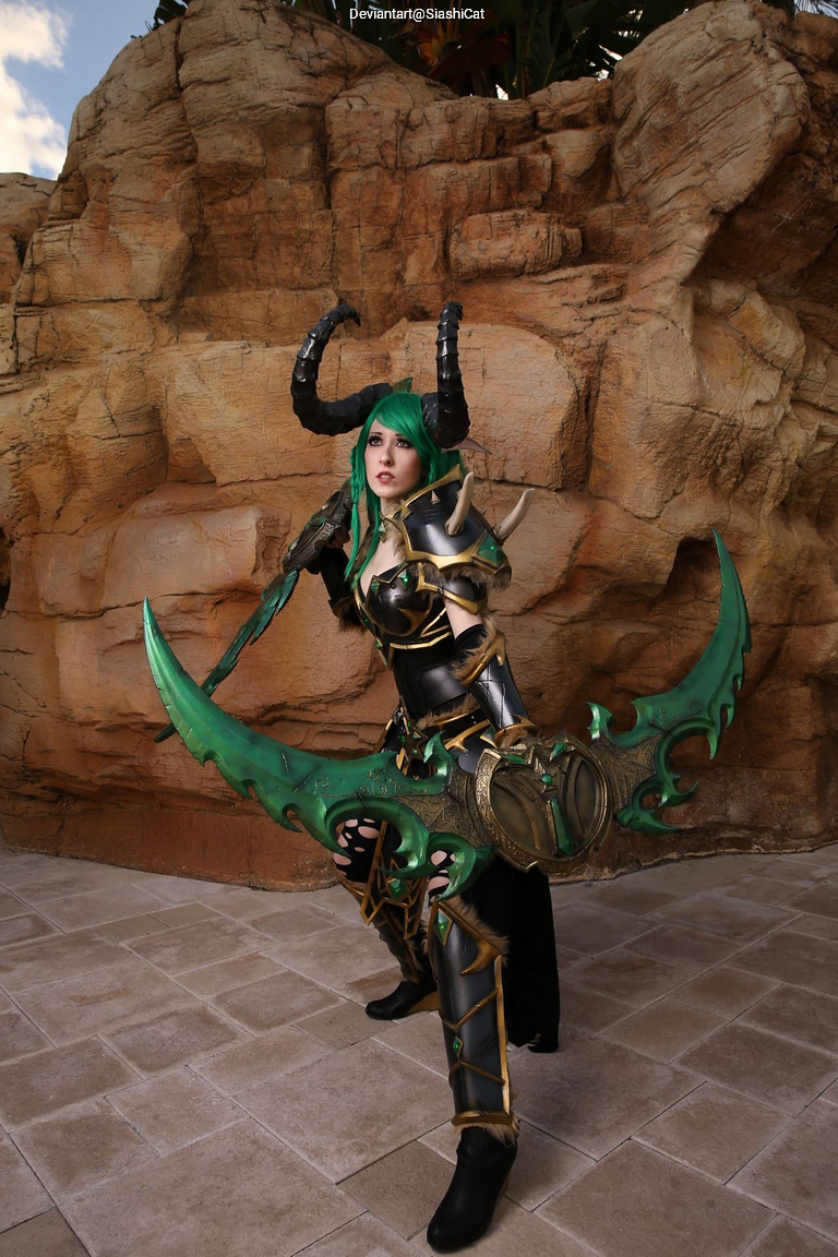 A cosplayer dressed in elaborate fantasy armor with green accents, wielding dual large weapons, poses confidently against a rocky backdrop.