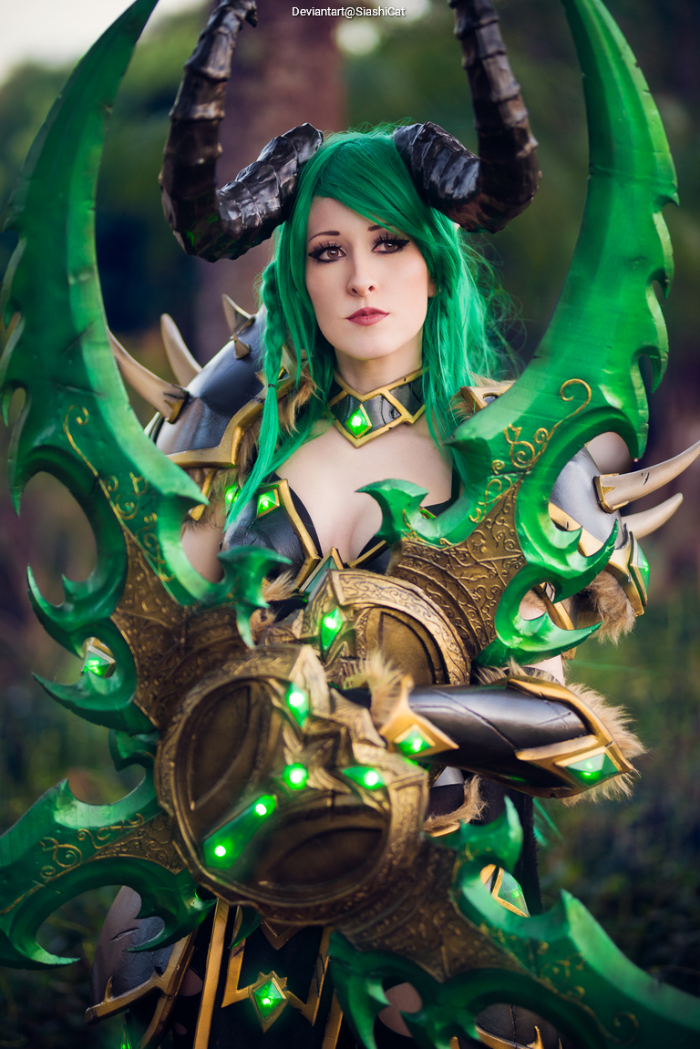 A person in elaborate fantasy costume with green hair and large, ornate horns. They are holding a pair of intricately designed, glowing green weapons, set against a natural background.