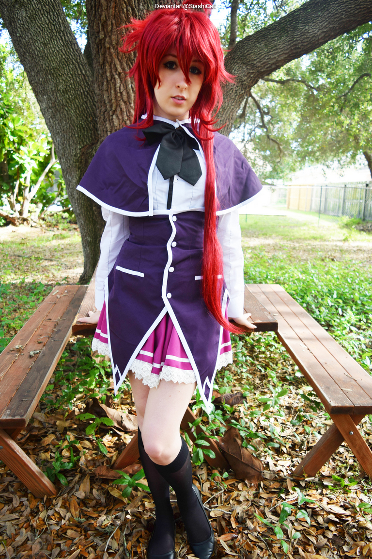 A person with long red hair dressed in a purple and white outfit stands in a park setting near wooden benches, surrounded by greenery and fallen leaves.
