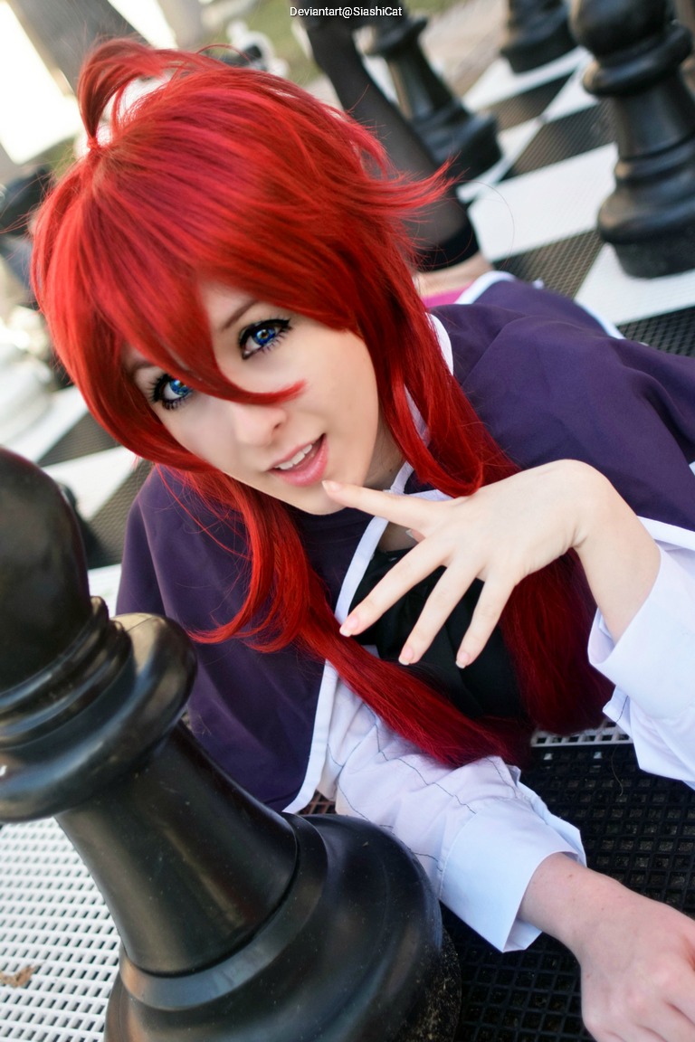 A person with bright red hair and striking blue eyes poses playfully next to a large black chess piece on a checkered board.