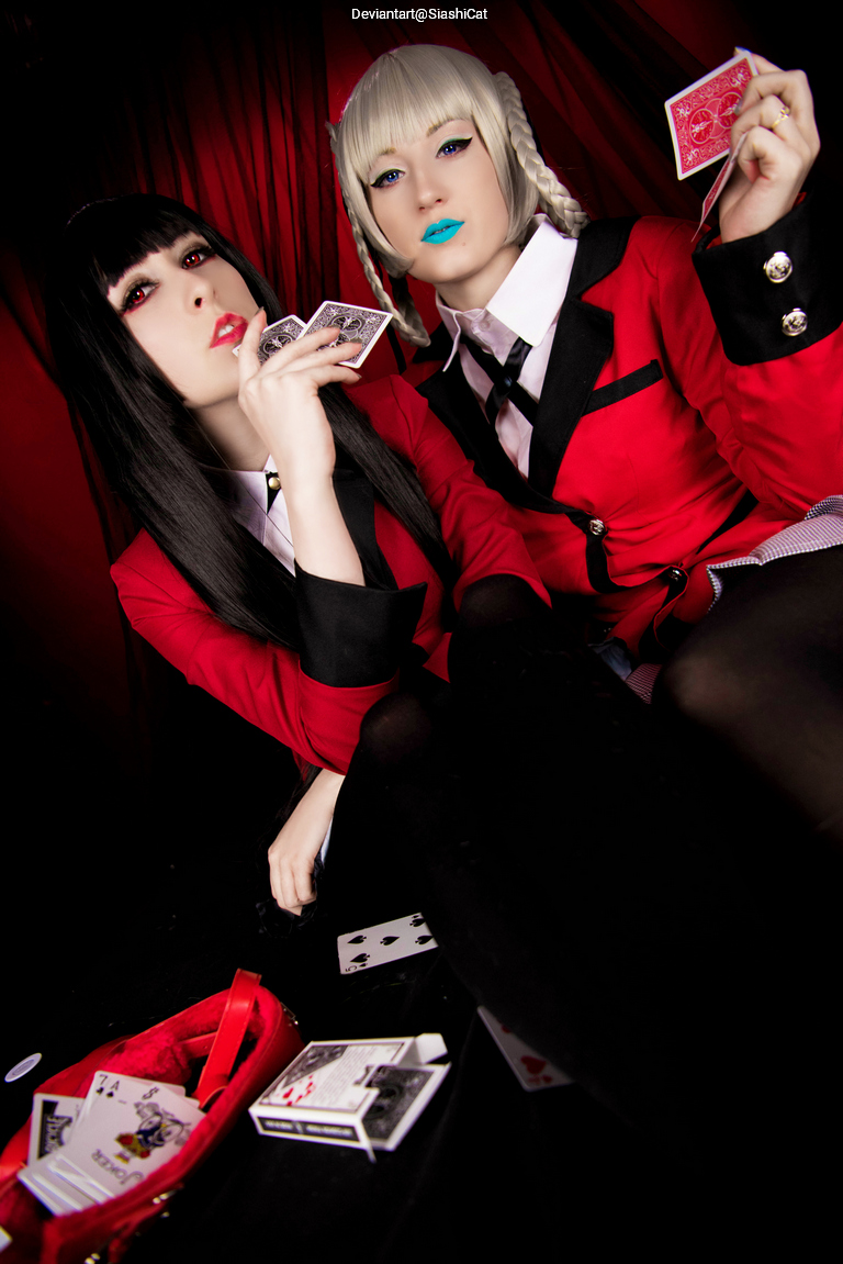 Two women in red blazers posing with playing cards, against a red backdrop. One has long dark hair and red lips, while the other has light hair and blue lipstick.