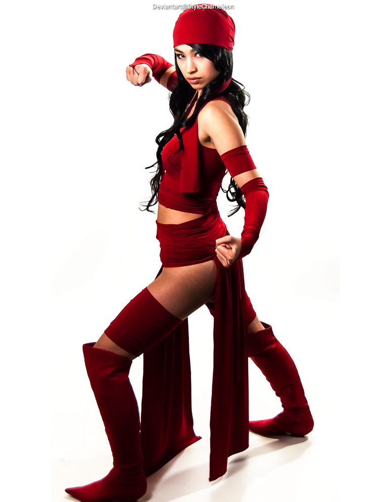 A woman poses in a dynamic stance, wearing a striking all-red outfit consisting of a cropped top, thigh-high socks, and a matching headwrap. Her long dark hair cascades over her shoulders as she appears ready for action.