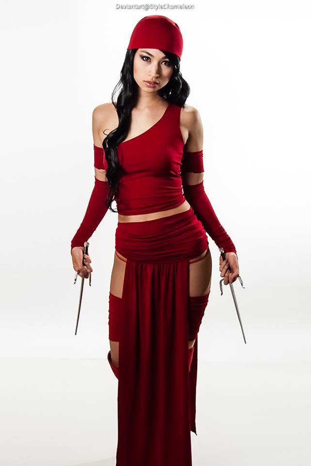 A woman dressed in a red outfit with cut-out details, holding two weapons, posing in front of a plain white background.