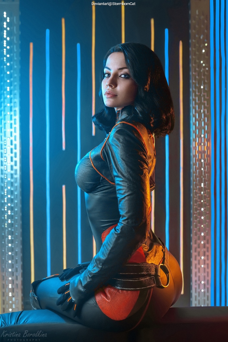 A woman wearing a form-fitting, black and red superhero costume poses with her back facing the camera, looking over her shoulder. The background features colorful vertical lights, enhancing the dramatic ambiance.