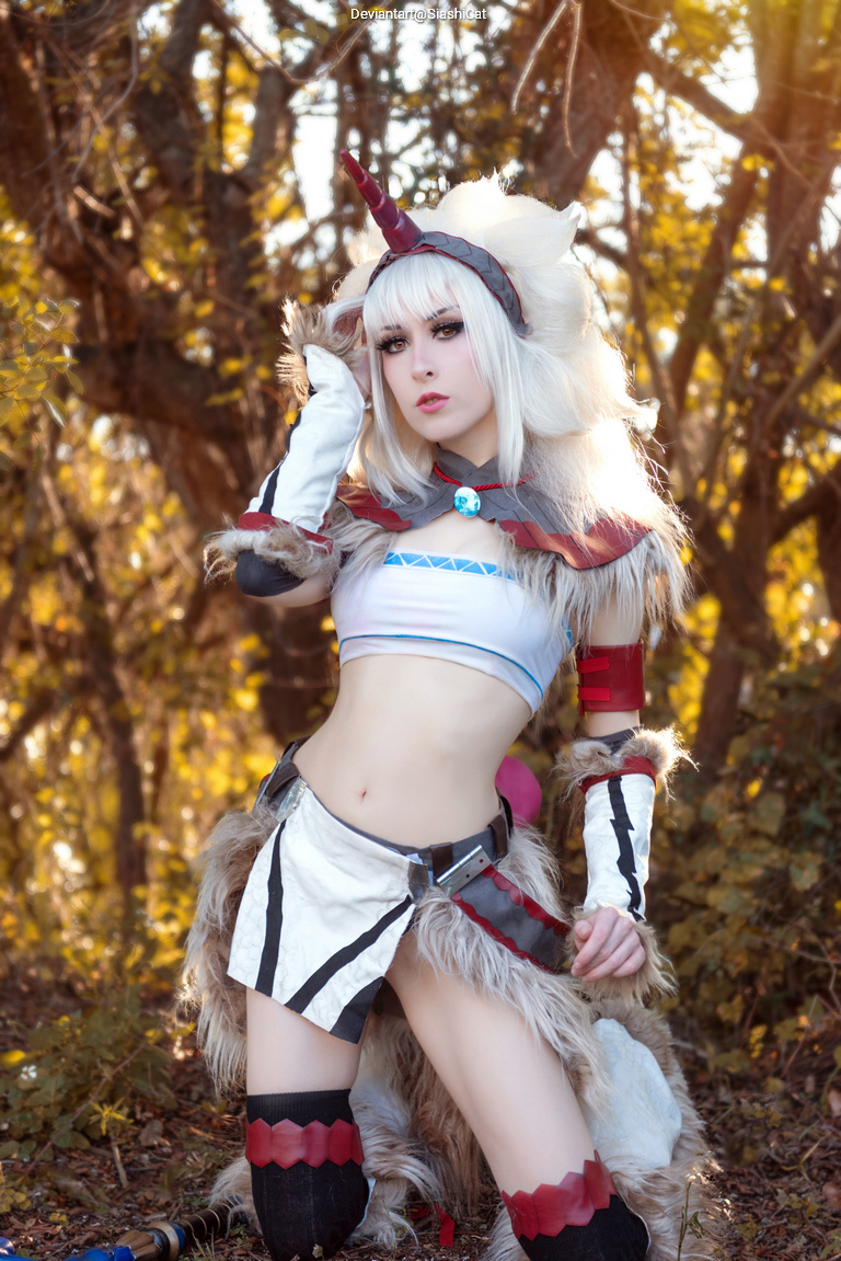 A posed shot of a female cosplayer dressed in a fantasy-inspired outfit, featuring a crop top and a skirt with fur accents. She has long blonde hair and a horned headpiece, set against a blurred background of trees with autumn foliage.
