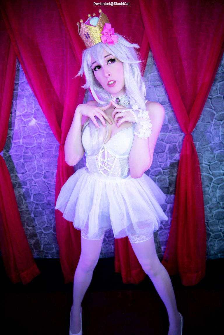 A person dressed in a white costume with a playful crown, standing in a theatrical pose in front of a purple and red backdrop.