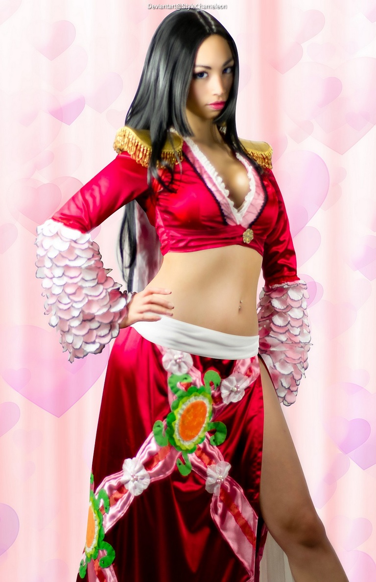 A woman in a vibrant red costume with floral embellishments, posing confidently against a soft pink heart-patterned background.