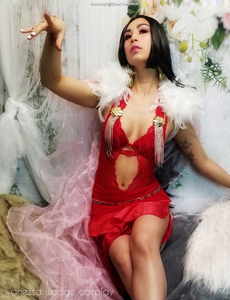 A woman in a striking red outfit with feathered accents, seated in a dramatic pose against a floral backdrop. She has long black hair and is adorned with elegant jewelry.