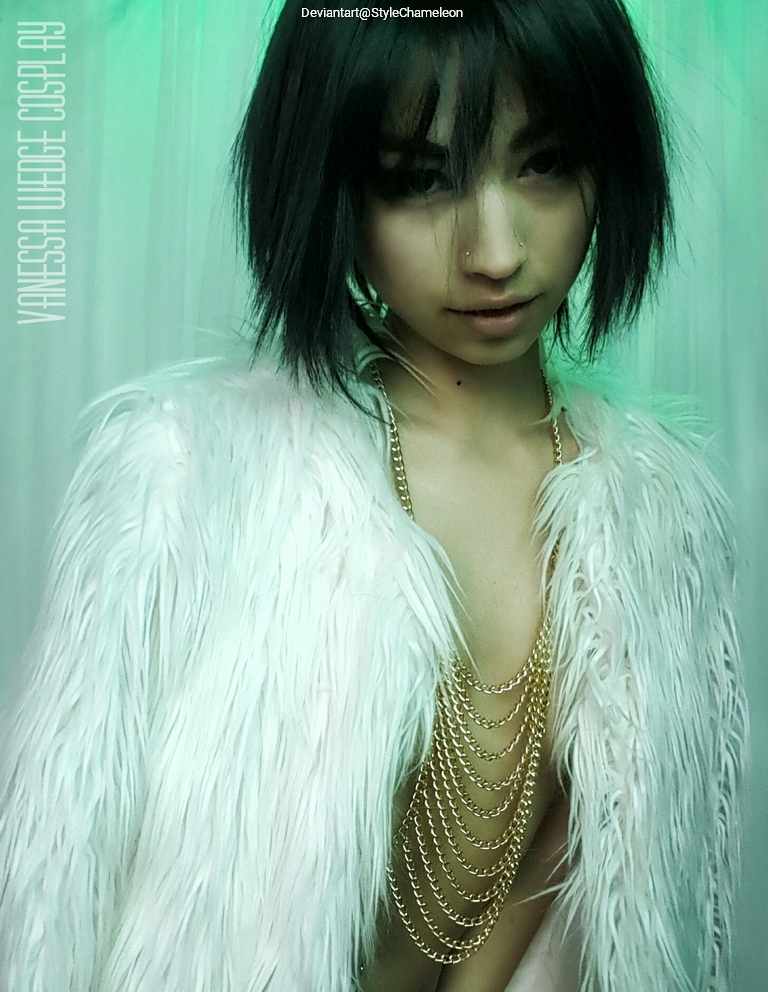 A person with short black hair wearing a white furry coat and layered gold chain jewelry, posing with a sultry expression against a green backdrop.