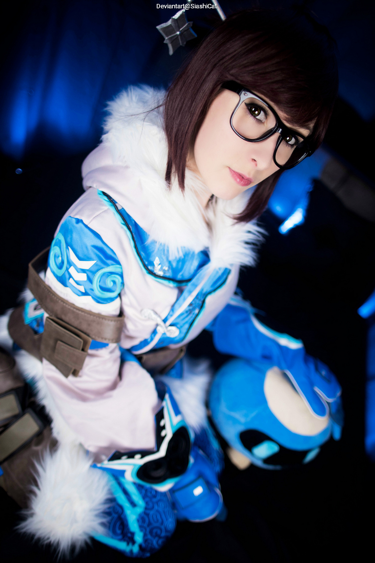 Cosplay of a character wearing a blue and white outfit with a fur-trimmed hood and glasses, holding a blue plush toy, set against a dark background with blue lighting.