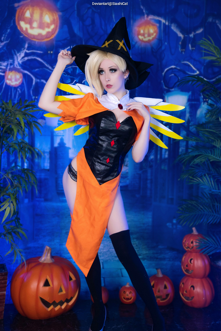 A person dressed in a witch costume with a black hat, orange and black clothing, standing in front of Halloween-themed decorations including pumpkins and a spooky backdrop.