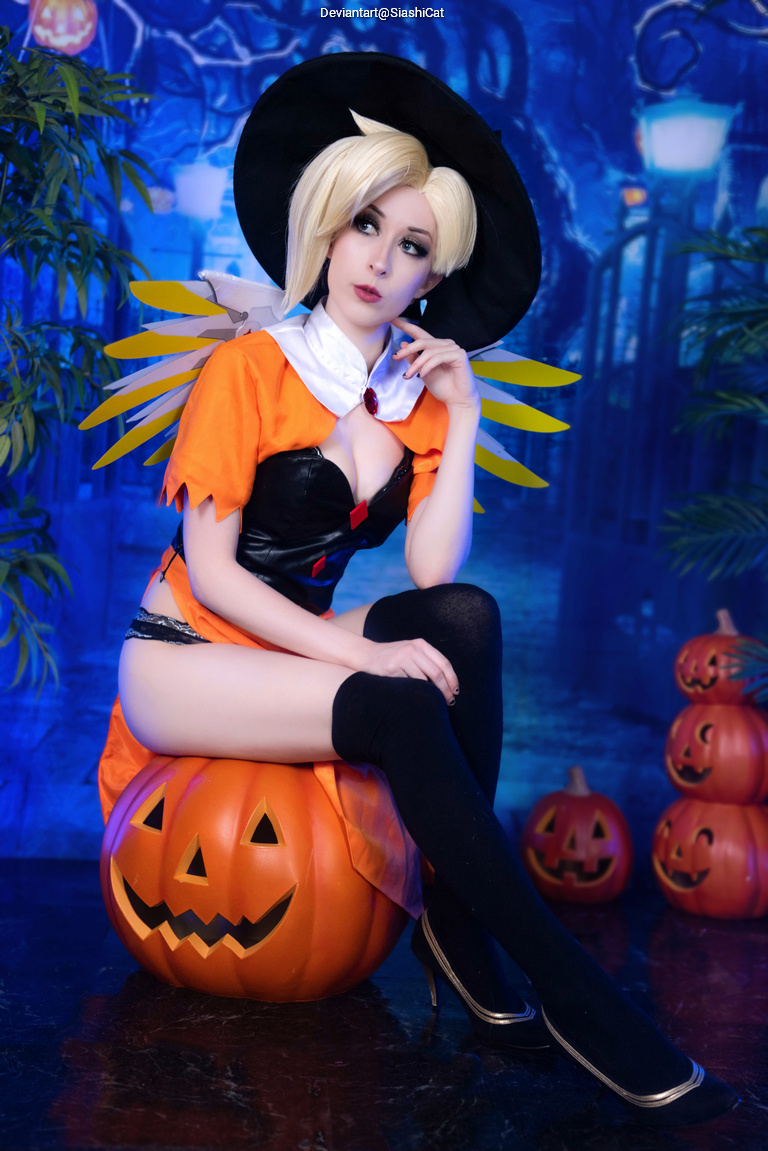A person in a witch costume, sitting on a carved pumpkin, surrounded by more pumpkins and a blue-hued background with Halloween decorations.