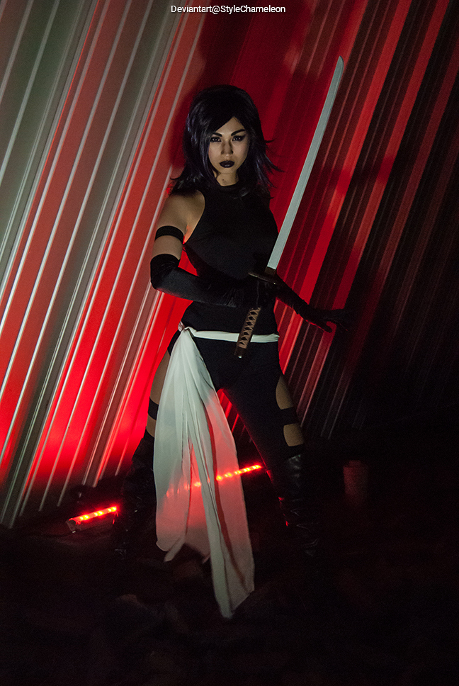 A person dressed in a dark, stylish costume holding a sword, posing dramatically in front of striped red and silver background lighting.