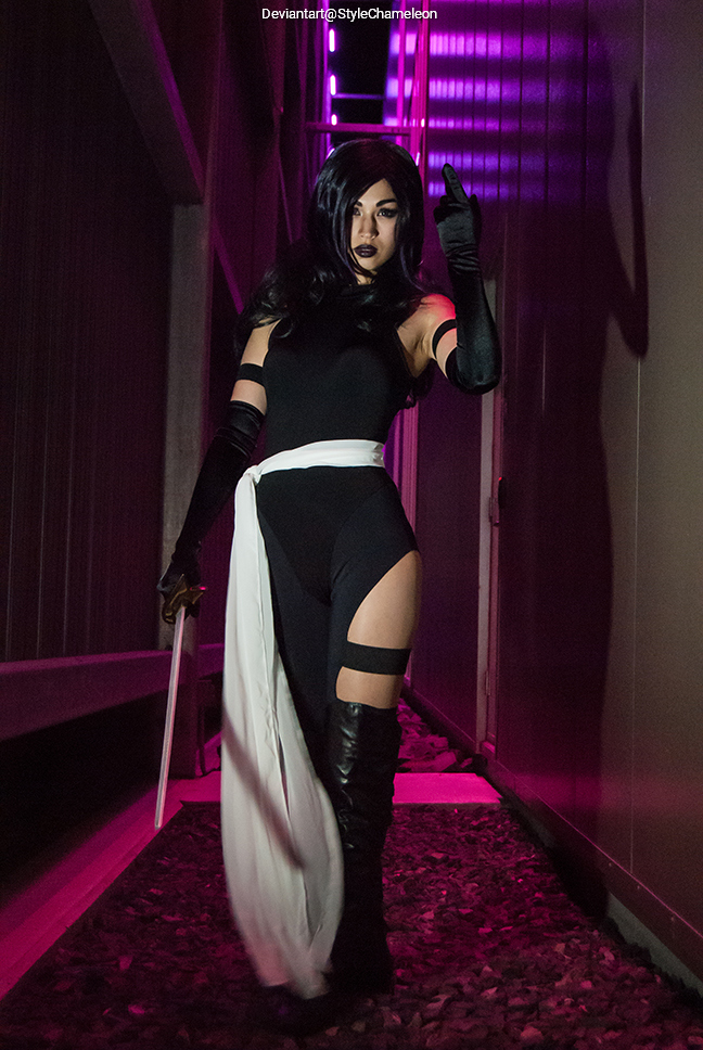 A person dressed in a black costume with a white sash, holding a sword, walking through a dimly lit path with purple lighting.