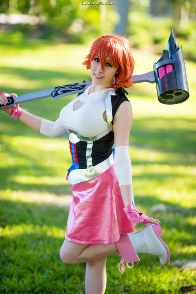 A person in a colorful costume with short red hair poses outdoors, holding a unique weapon, amidst a green grassy background.