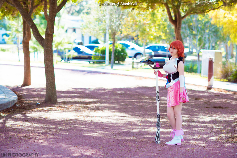 A person wearing a pink costume stands in a park, holding a large staff, with trees and parked cars in the background.