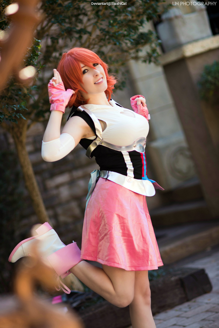 A person with short orange hair wearing a colorful outfit, striking a playful pose with one leg raised and a cheerful smile, surrounded by greenery.