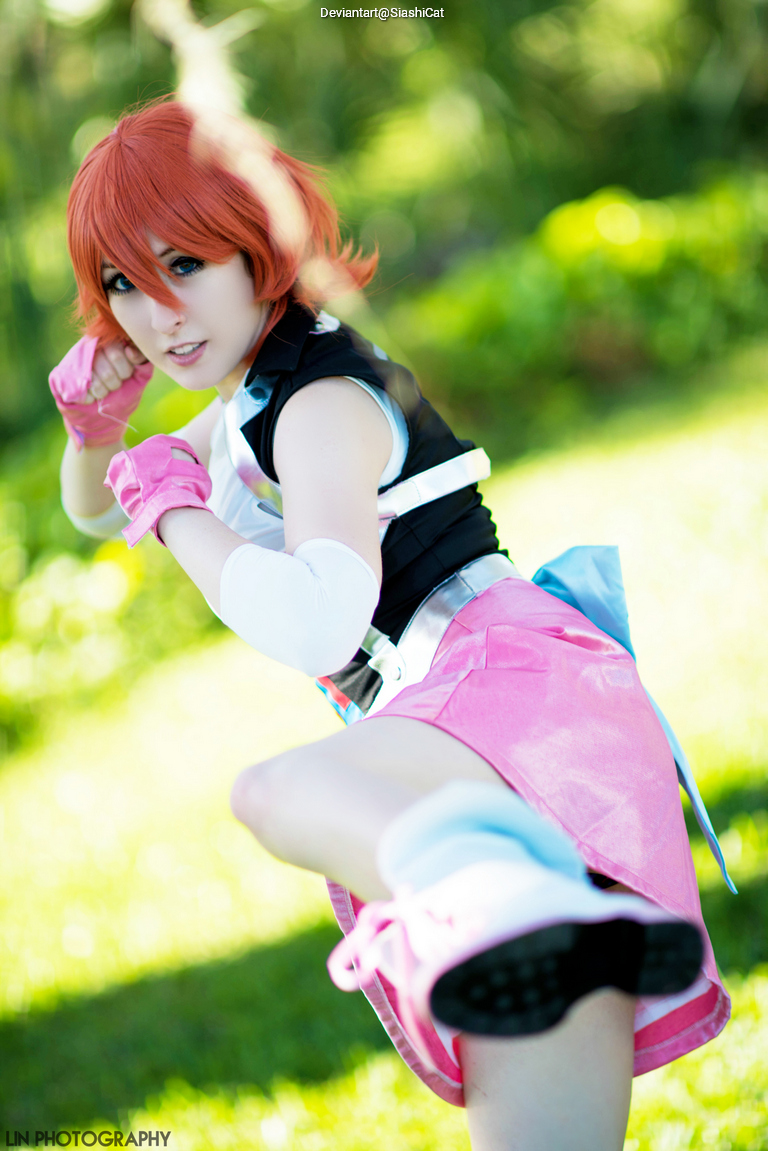 A person in a costume performs a dynamic kick, wearing a pink skirt and a black top, with short orange hair and gloves, set against a blurred green background.