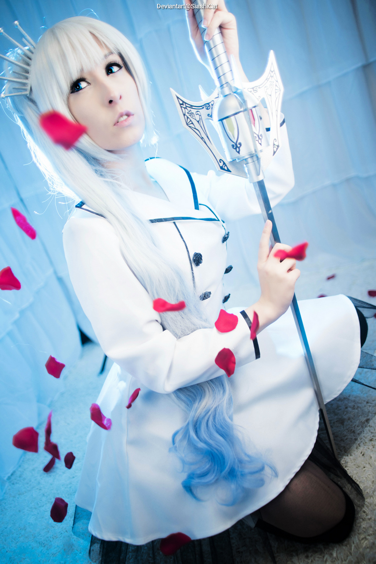 A person in a white outfit with a long, silver wig sits on the floor, holding a sword, surrounded by rose petals. The background is soft-focused and light-colored.