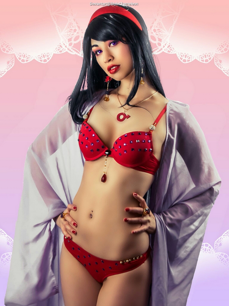 A woman posing confidently in a red bikini adorned with jewels, wearing a matching robe and accessories, against a soft pink background.