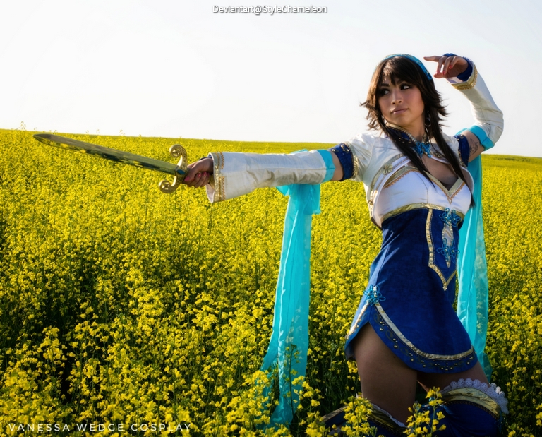 A female cosplayer dressed in blue and white attire, holding a sword, poses dramatically in a field of yellow flowers.