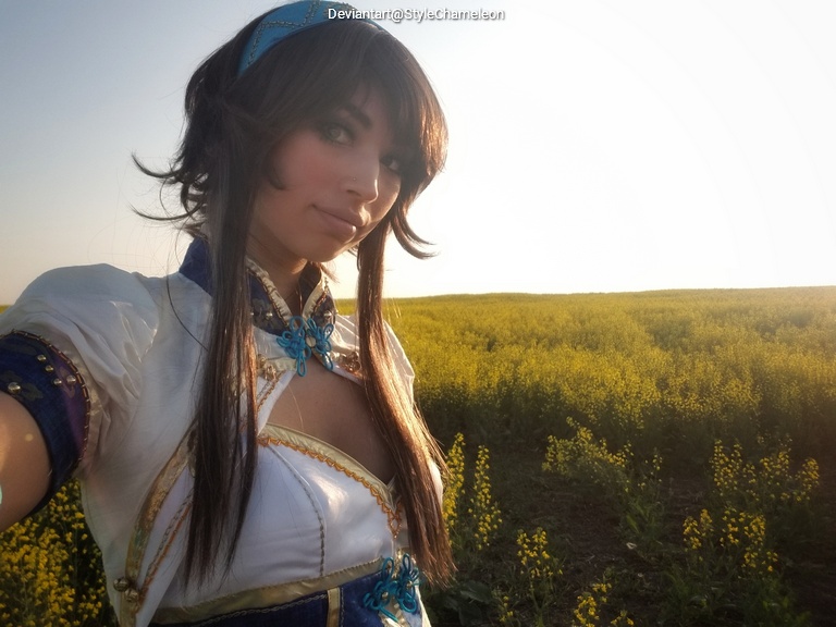 A woman with long hair wearing a traditional-inspired costume with blue and white accents poses for a selfie in a yellow flower field during sunset.