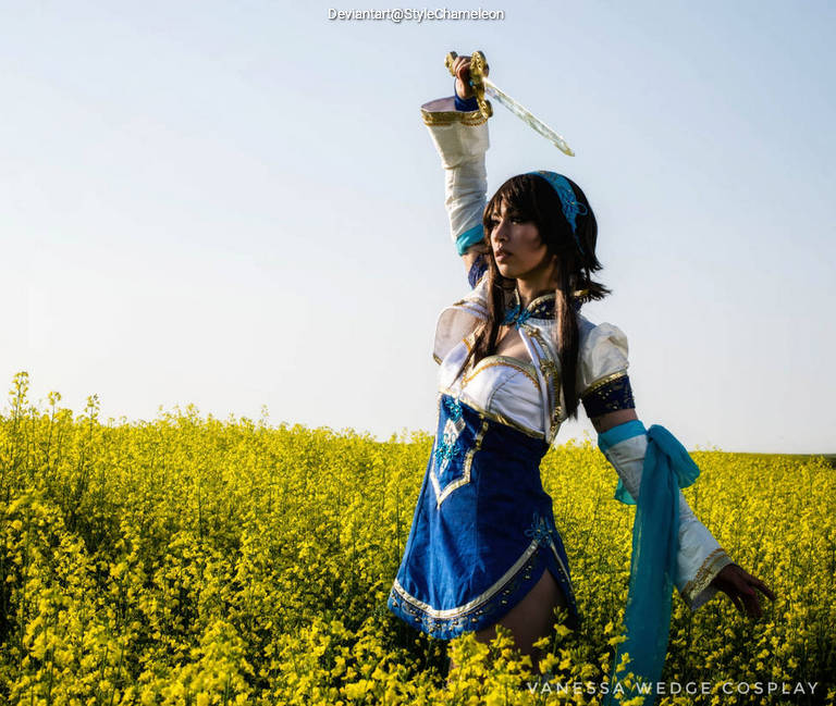 A female cosplayer dressed in a blue and white outfit, wielding a sword, standing in a field of yellow flowers.