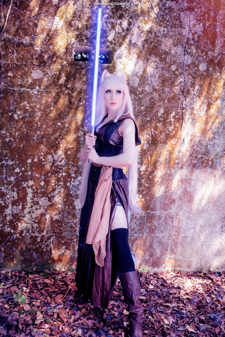 A person dressed in a character costume with long blonde hair, holding a glowing blue lightsaber in a forest setting with autumn leaves.