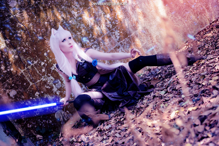 A cosplayer in a dark outfit with long blonde hair and blue accessories poses dramatically in a forest setting, holding a blue lightsaber.