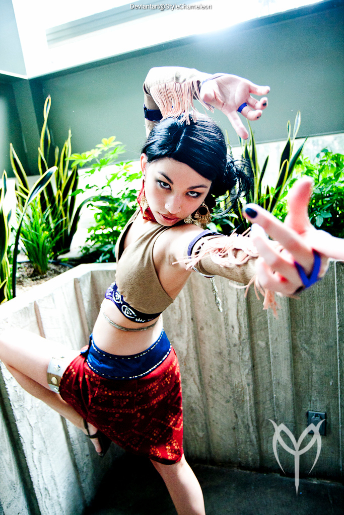 A woman in a dynamic pose, showcasing a costume with fringe details and colorful accessories, set against a backdrop of greenery and concrete.