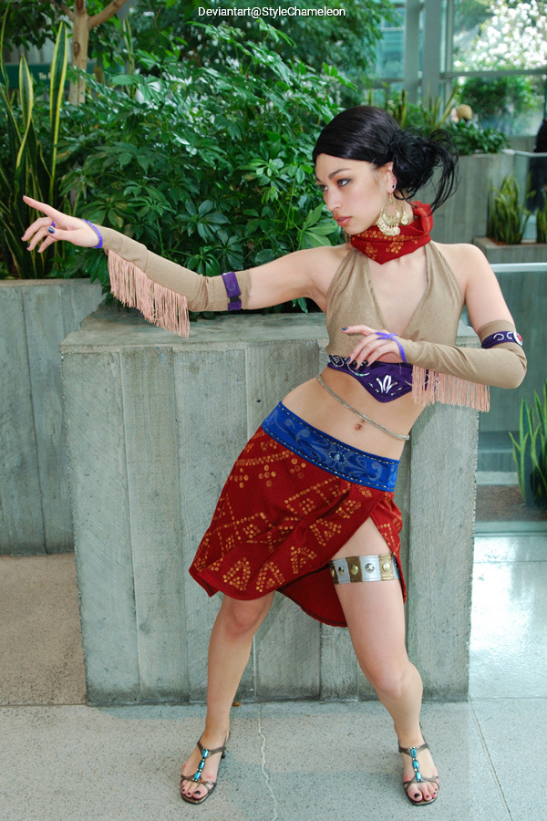 A person in a costume striking a dynamic pose with one arm extended, wearing a beige top, a red skirt with patterns, and colorful accessories in a green indoor setting.