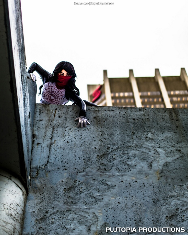 A person dressed as a superhero with a spider web design on their suit and a red mask, perched on the edge of a concrete ledge, looking down.