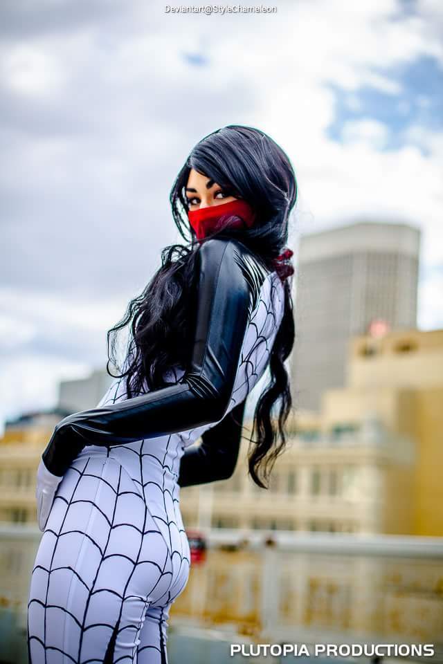 A person in a spider-themed costume with a black and white design, red mask, and long dark hair, standing outdoors against a cloudy sky.