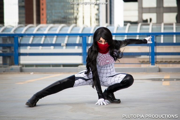 A person dressed as a superhero in a white and black spider-themed costume with a red mask, striking a dynamic pose on a rooftop with city buildings in the background.