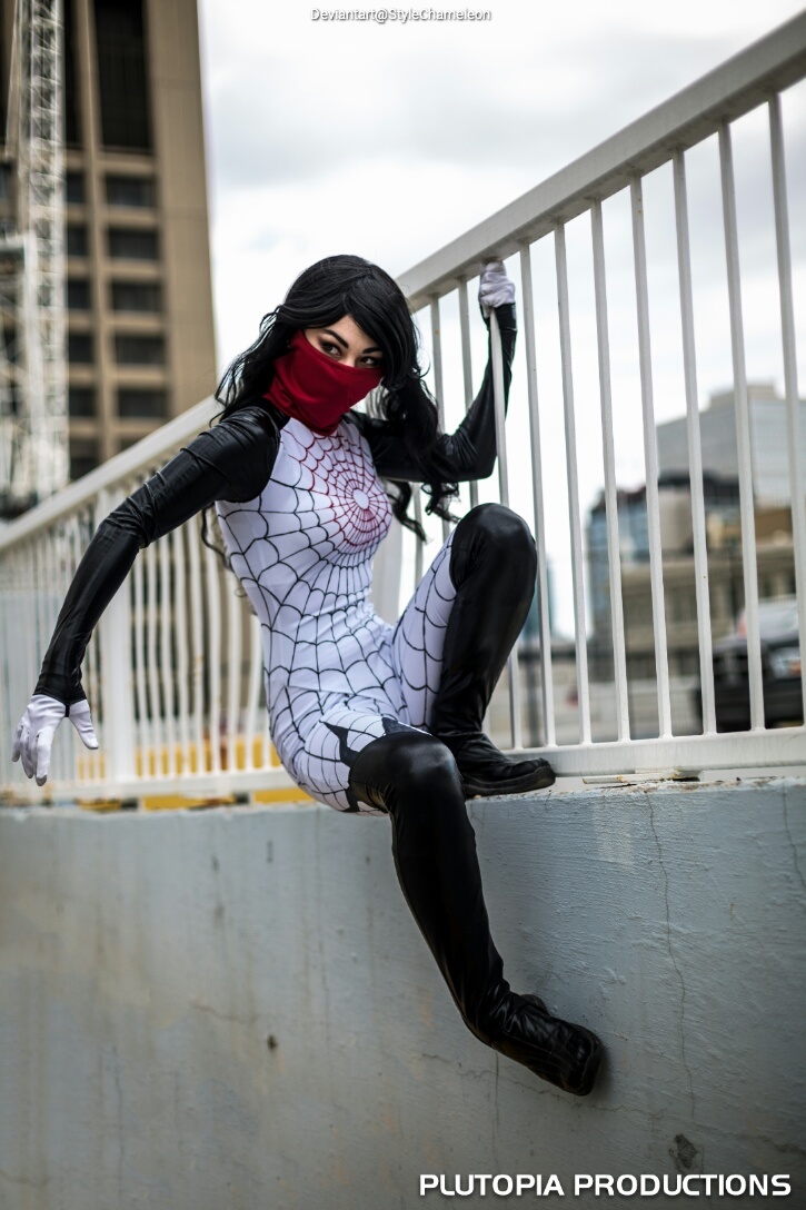 A person dressed in a spider-themed costume with a red mask and web patterns, crouched on a ledge in a city setting.