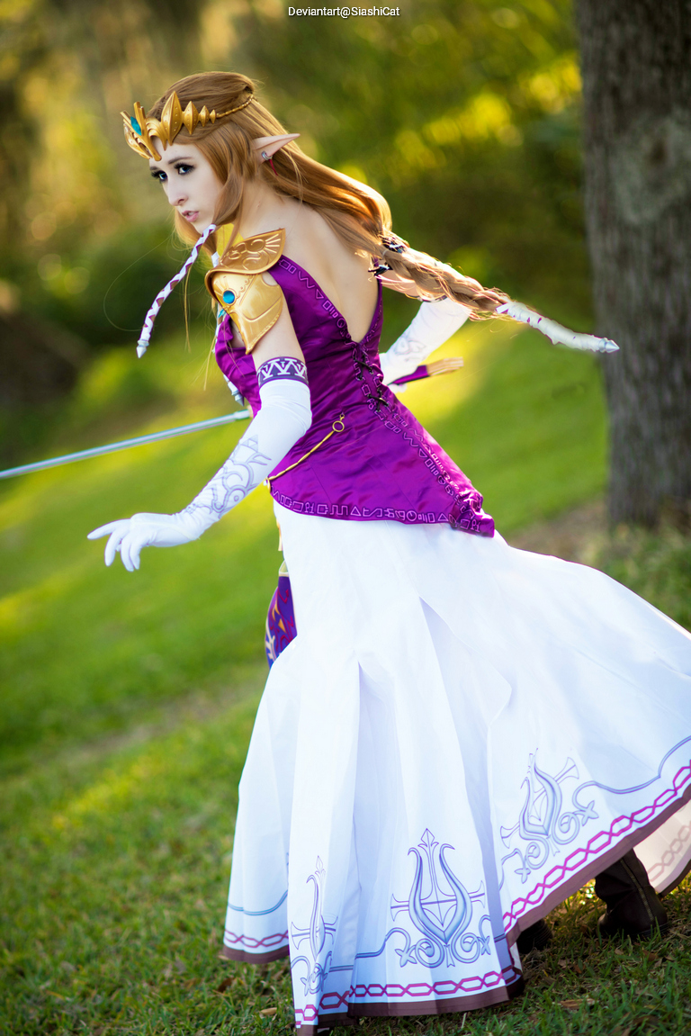 A person dressed as a princess with long blonde hair, wearing a purple and white costume, poses confidently outdoors with a blurred green background.