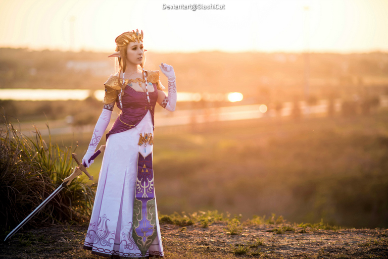 A person in elaborate fantasy costume resembling a princess, standing with a sword at sunset.