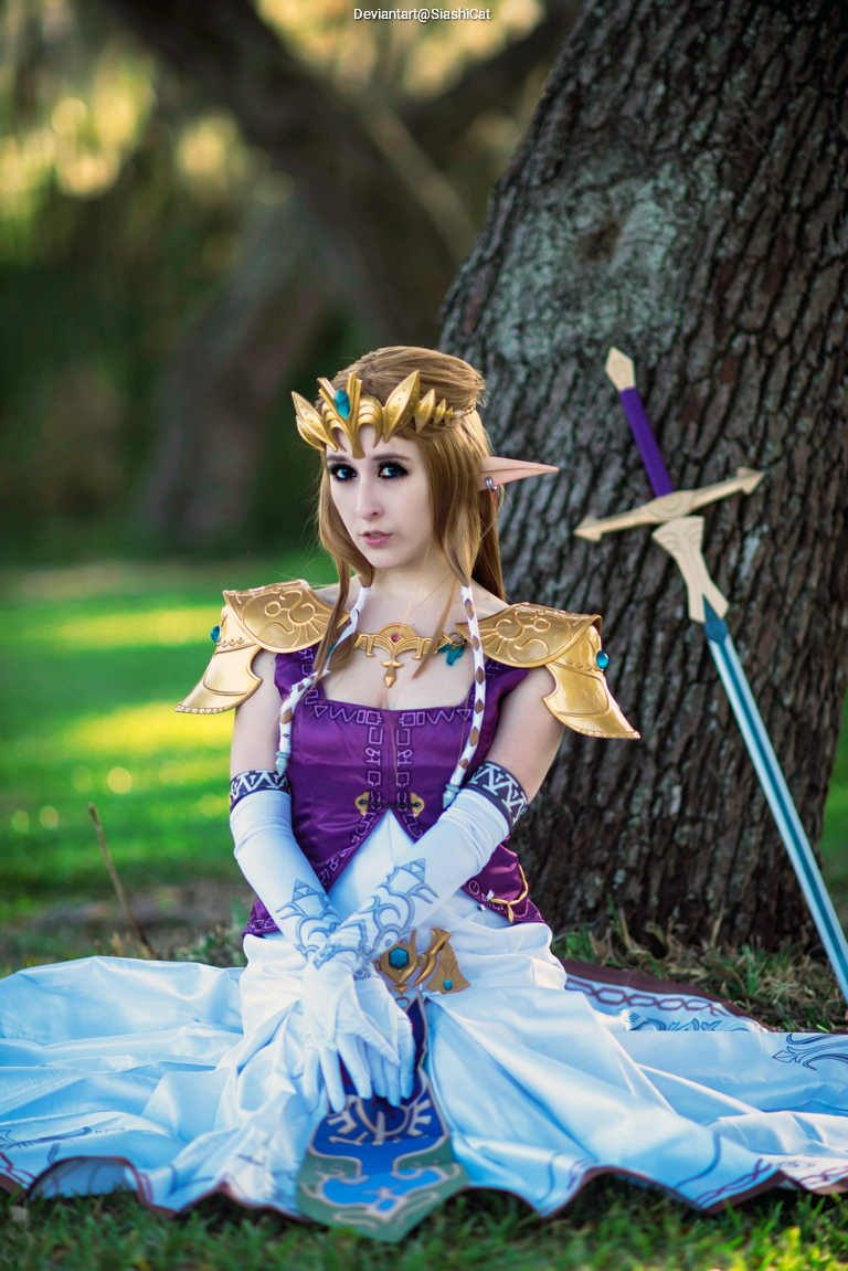A person dressed as Princess Zelda from the Legend of Zelda series, sitting on the ground in a natural setting, wearing a purple and white costume with a crown and a sword nearby.