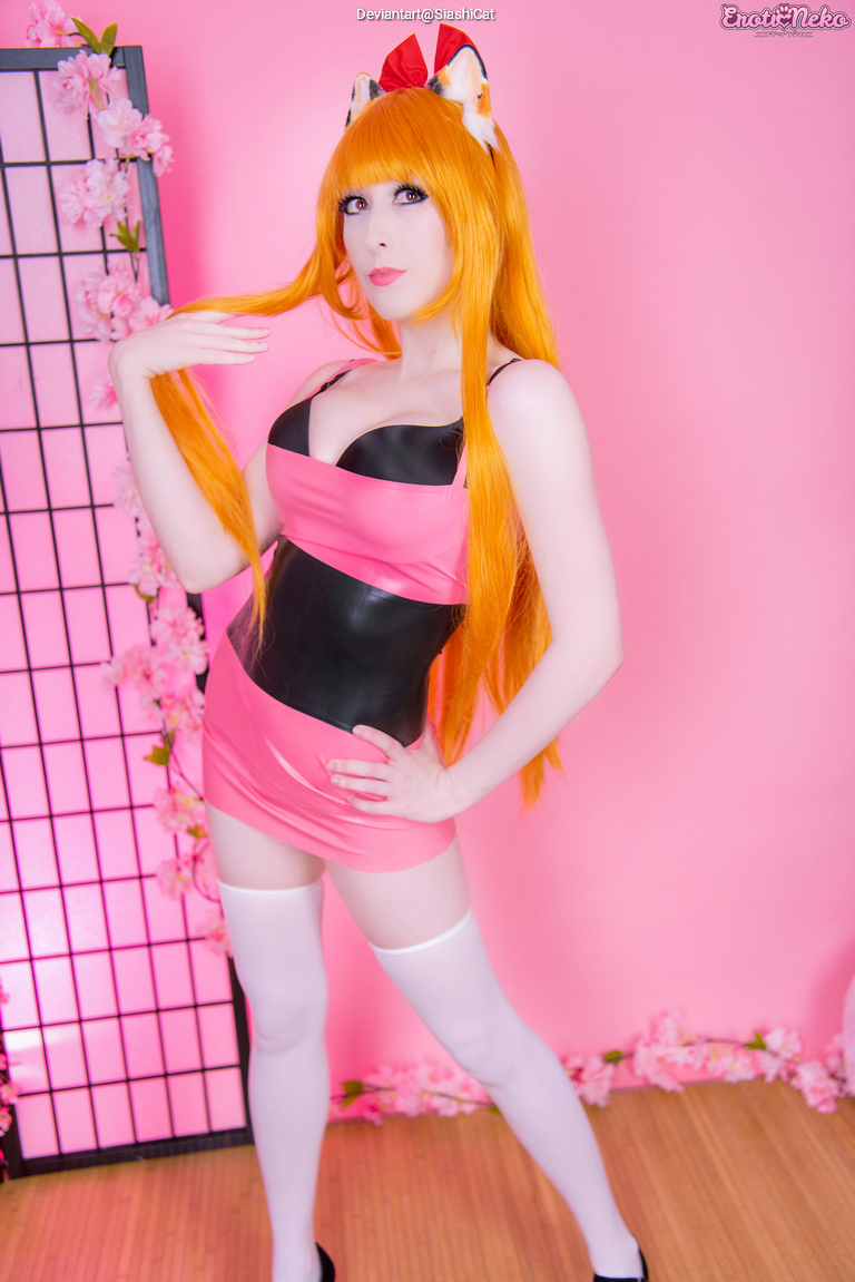 A person posing in a pink and black outfit with orange hair, styled in a referential fashion to a character, against a pink backdrop with decorative cherry blossom elements.