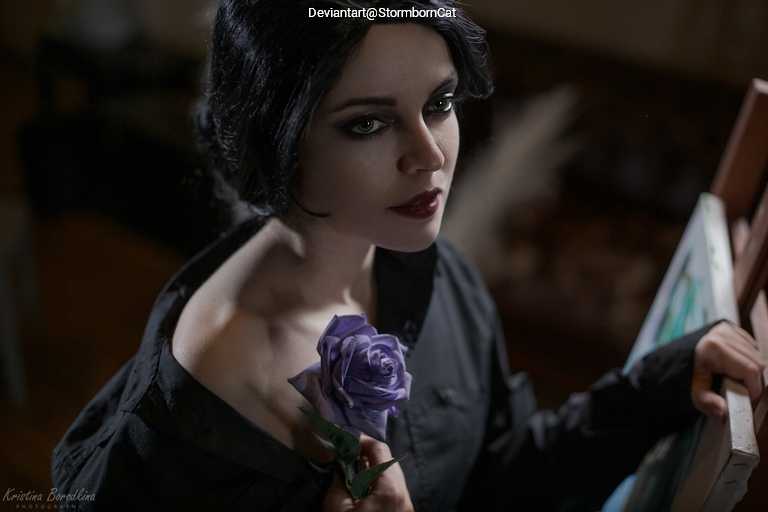 A woman with dark hair and dramatic makeup holds a purple rose, staring thoughtfully off-camera while positioned next to an easel with a painting.