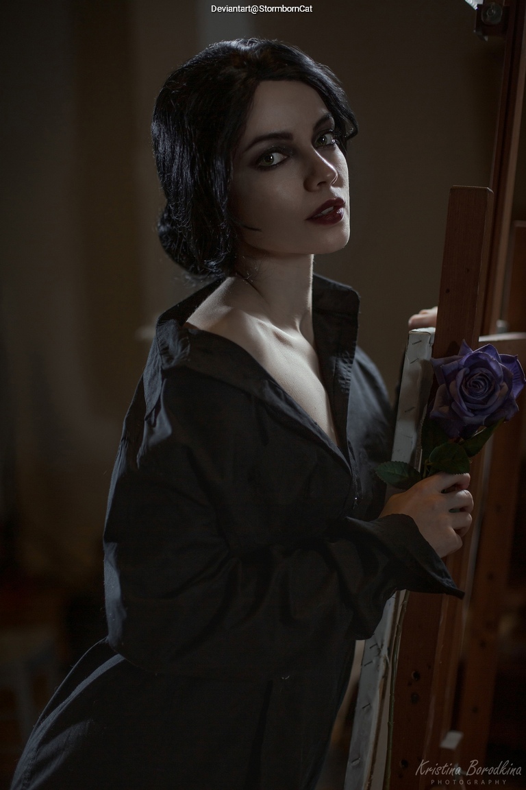 A woman with dark hair and striking makeup, wearing a black shirt, poses holding a purple rose in front of an easel in a softly lit room.