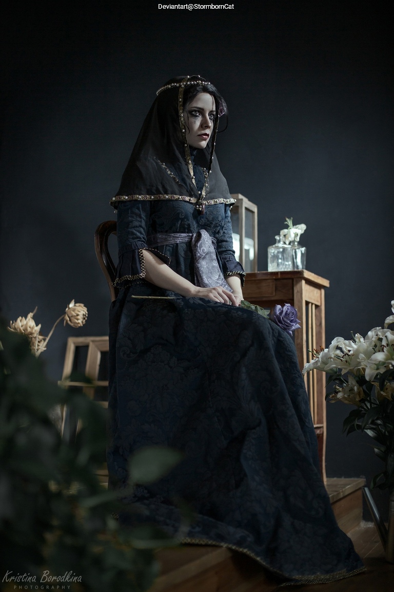 A woman in a dark, elegant dress and a headscarf sits on a wooden chair, surrounded by flowers and antique decor in a moody setting.