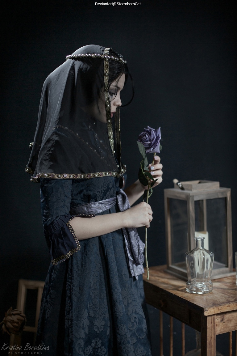 A woman in a dark vintage dress and veil holding a purple rose, standing beside a wooden table with a lantern and a glass bottle, against a dark background.