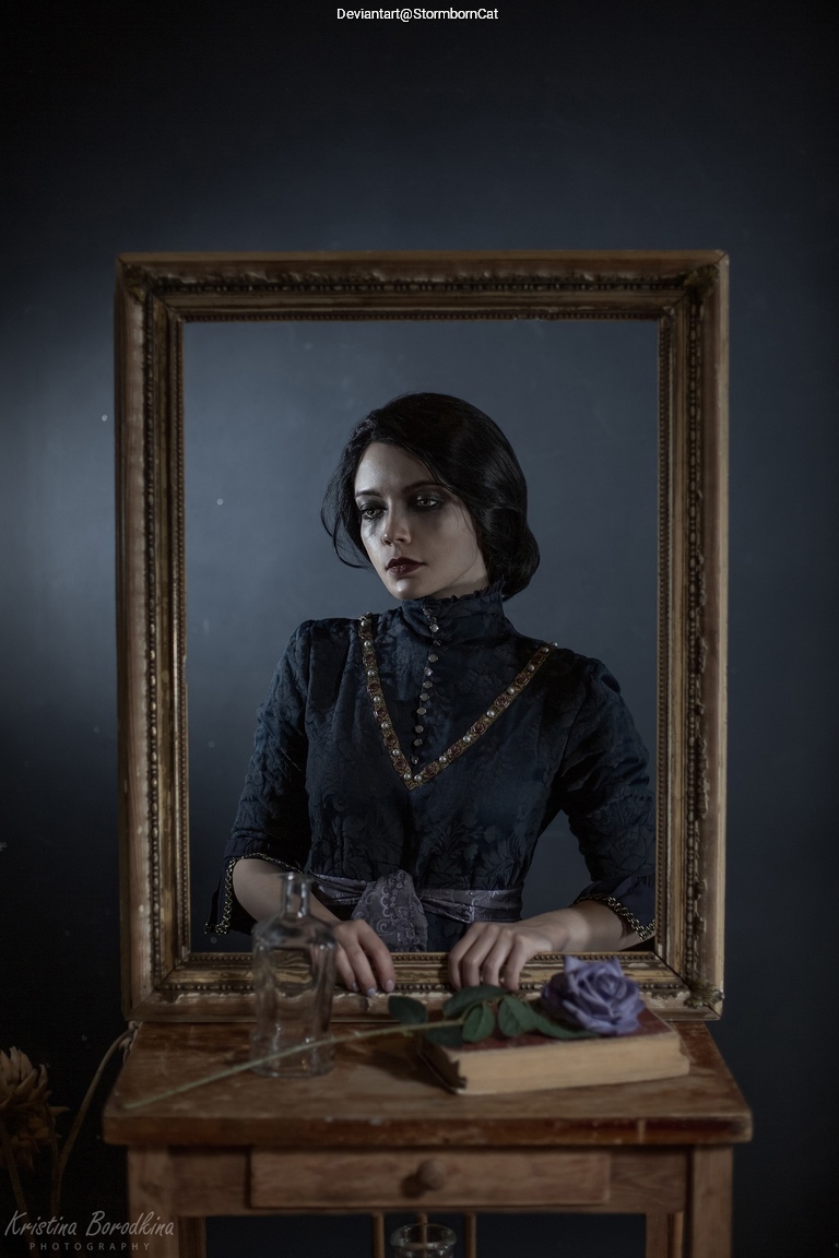 A woman in a vintage black dress posing in front of an ornate mirror, with a table displaying a book, a glass bottle, and a purple rose.